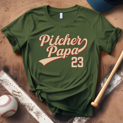 Green t-shirt with 'Pitcher Papa 23' text on a wooden surface with baseball equipment.